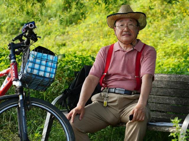 Un homme âgé en chapeau de paille, assis sur un banc dans la verdure, son vélo à panier posé à côté de lui.