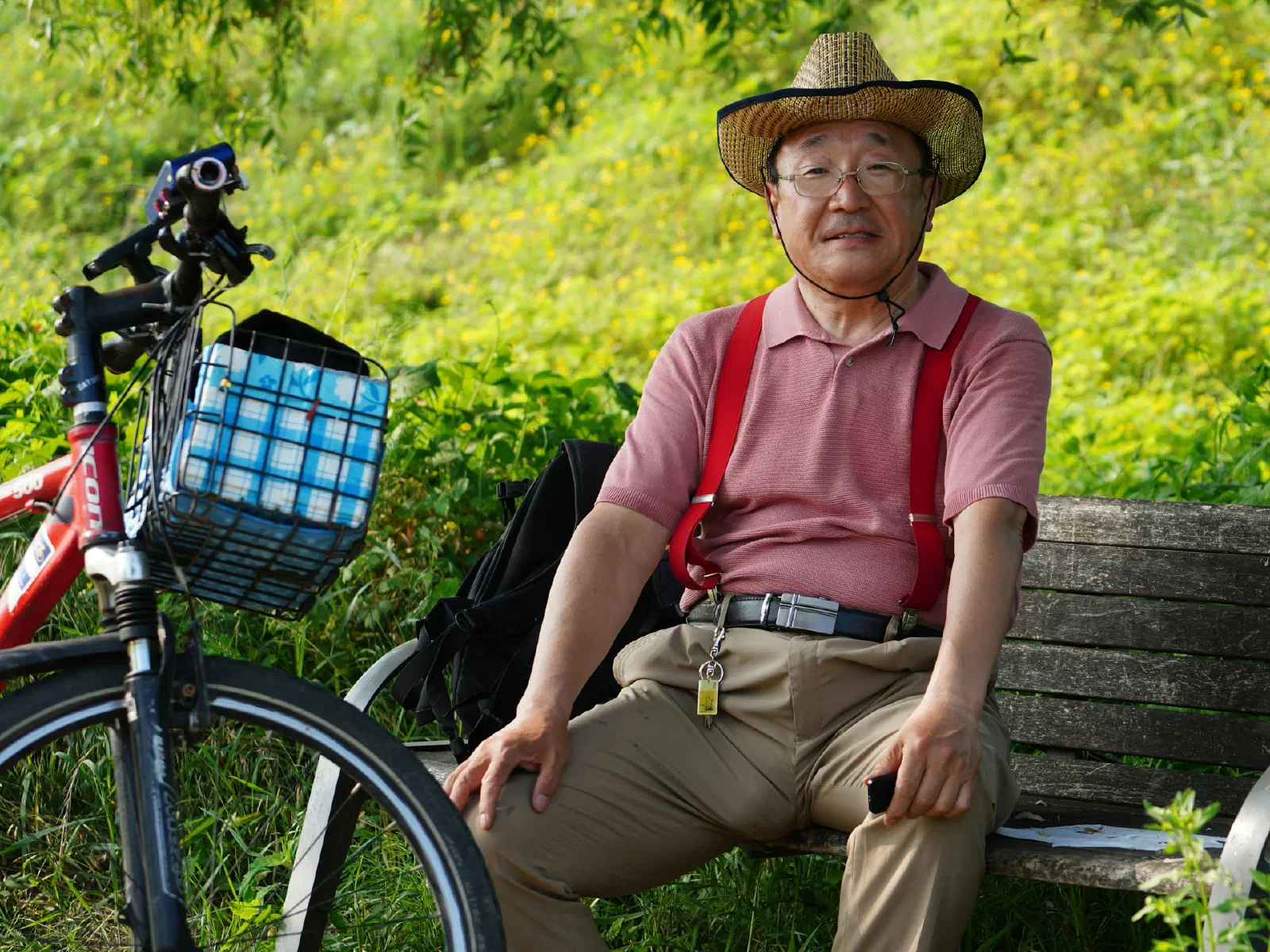 Un homme âgé en chapeau de paille, assis sur un banc dans la verdure, son vélo à panier posé à côté de lui.