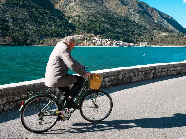 Un homme aux cheveux gris roule sur un vélo à panier le long d'une promenade en bord de mer, montagnes en arrière-plan.