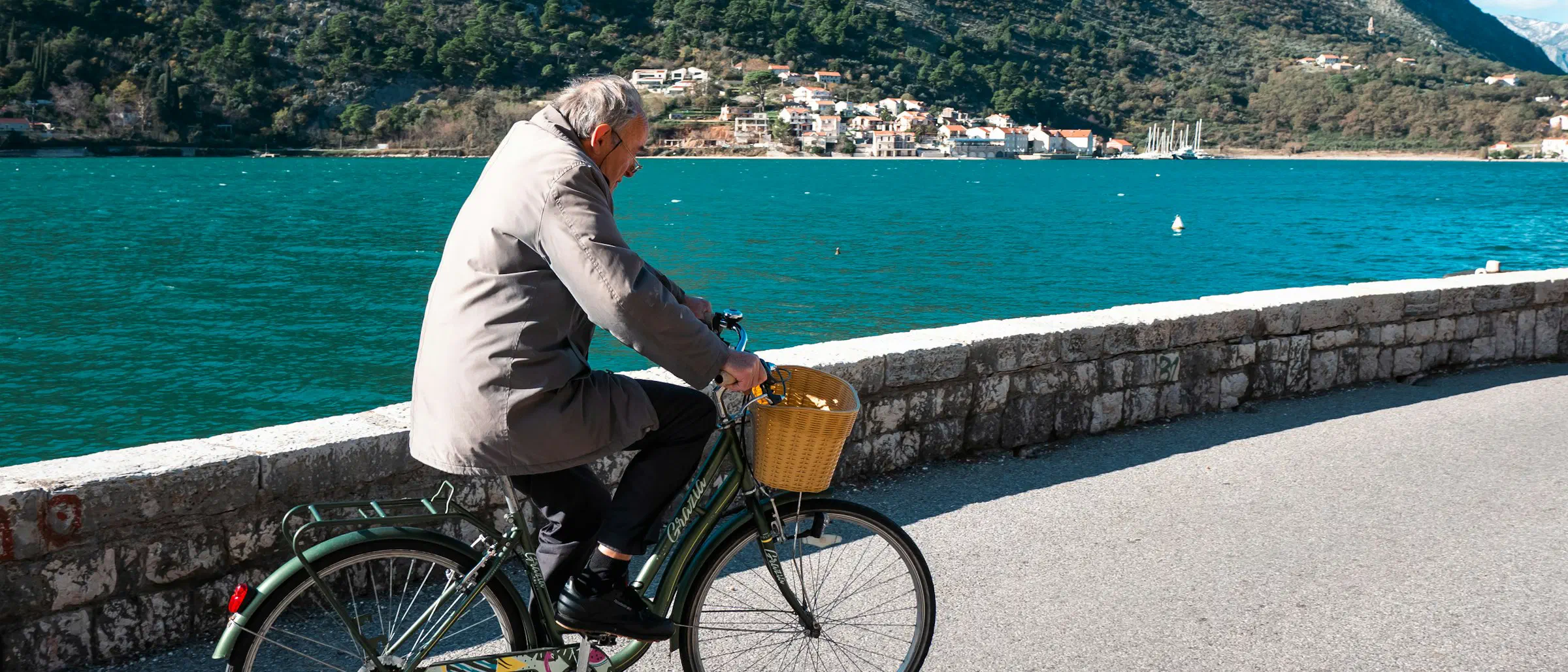 Un homme aux cheveux gris roule sur un vélo à panier le long d'une promenade en bord de mer, montagnes en arrière-plan.