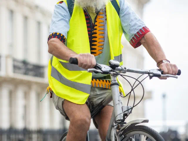 Un homme à barbe blanche, avec casque et gilet jaune de haute visibilité, roule dans une rue en ville.