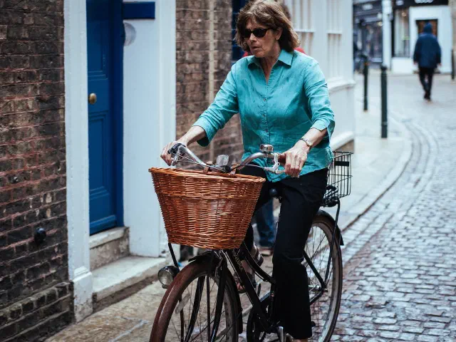 Une femme aux cheveux bruns, chemise turquoise, roule sur un vélo à panier en osier dans une ruelle pavée bordée de maisons en brique.