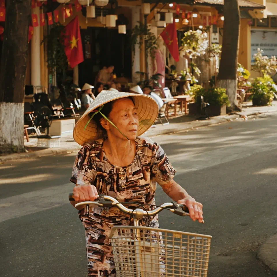 Une femme âgée coiffée d'un chapeau conique roule à vélo dans une rue animée, entre des scooters garés.