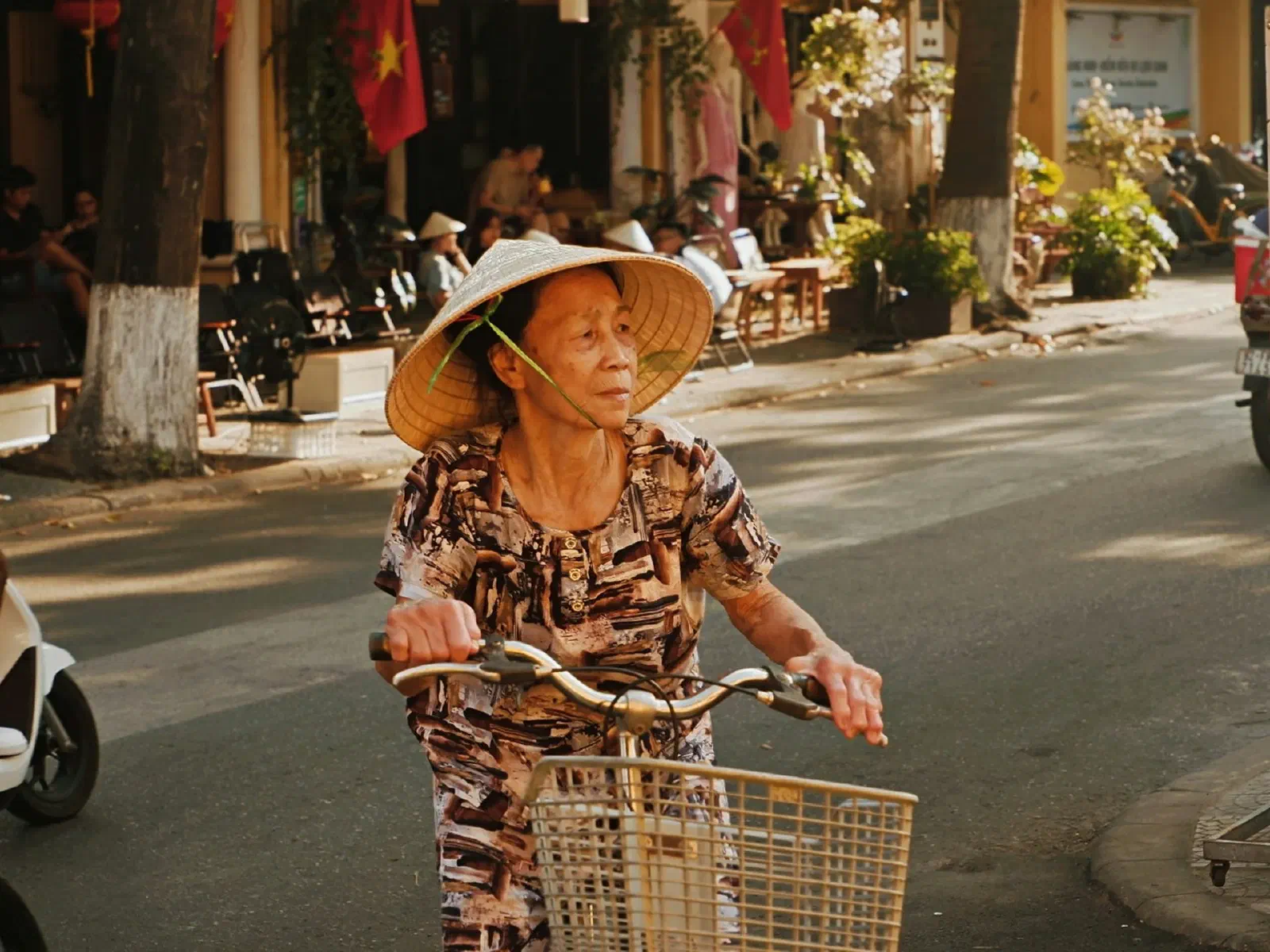 Une femme âgée coiffée d'un chapeau conique roule à vélo dans une rue animée, entre des scooters garés.