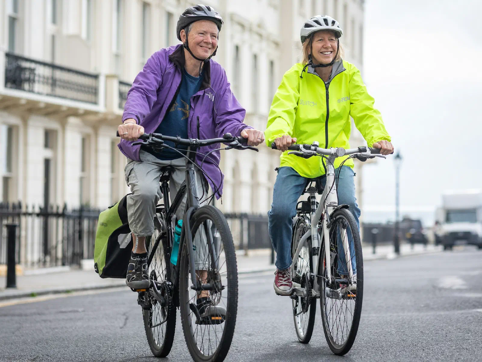 Deux cyclistes casquées, l'une en veste violette, l'autre en veste jaune fluo, roulent côte à côte dans une rue bordée d'immeubles blancs.