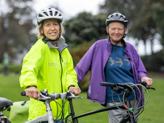 Deux femmes souriantes, casque sur la tête, debout à côté de leurs vélos dans un parc.