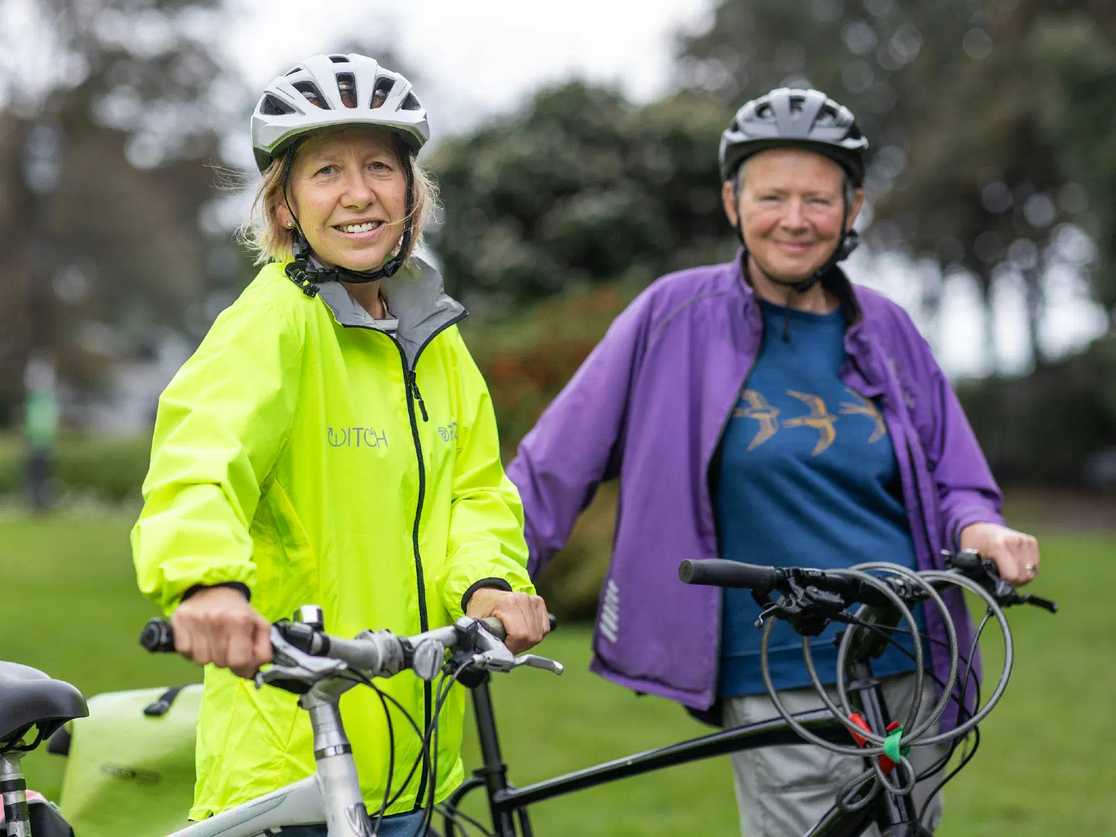 Deux femmes souriantes, casque sur la tête, debout à côté de leurs vélos dans un parc.