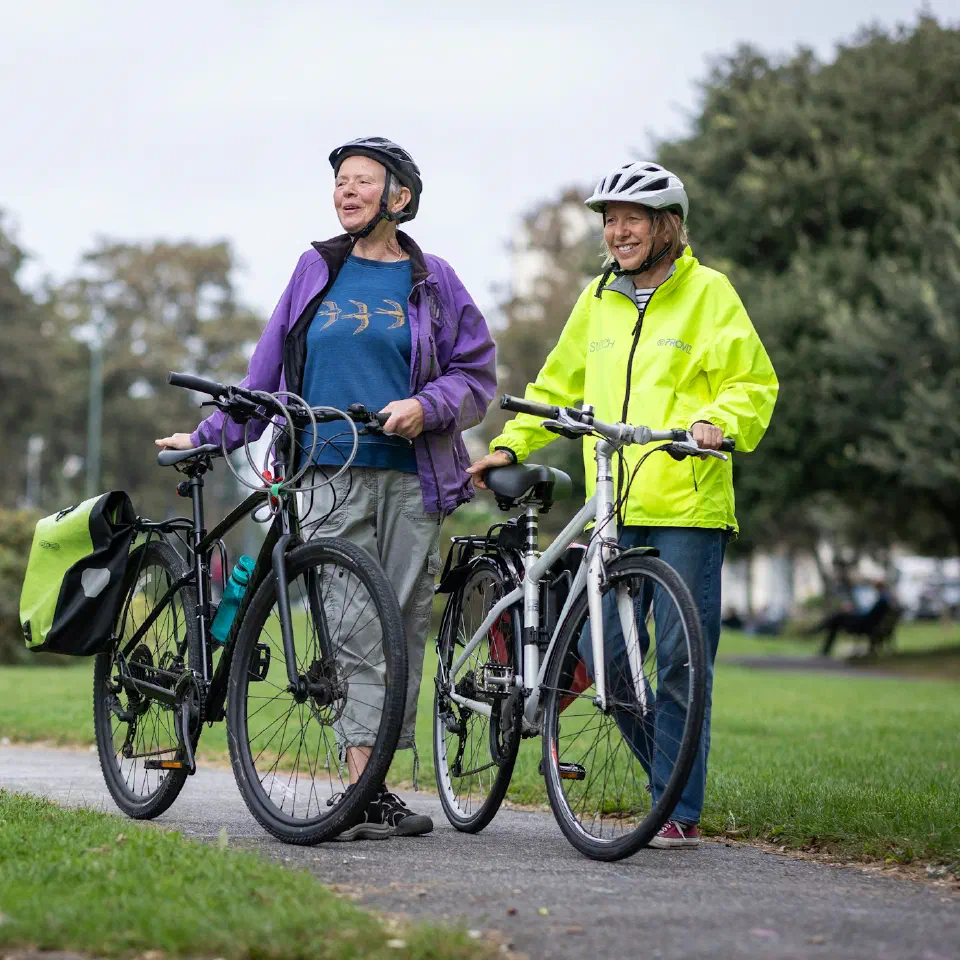 Deux femmes casquées poussent leurs vélos à la main sur une allée de parc.