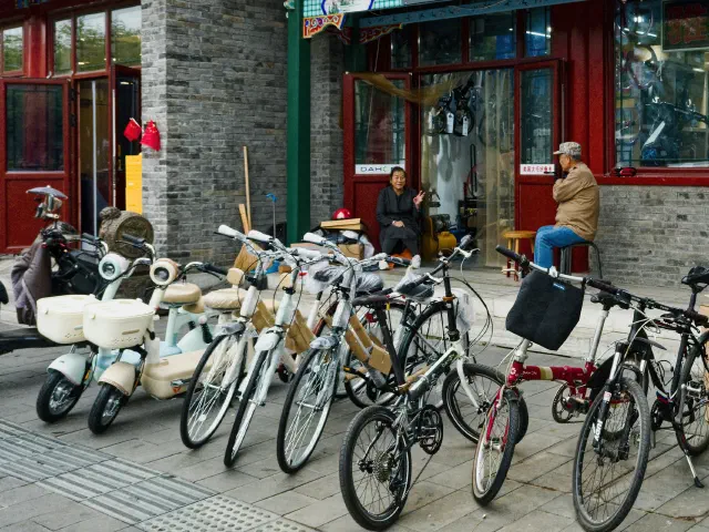 Vélos et scooters électriques alignés devant un magasin de cycles, deux personnes âgées assises à l'entrée.