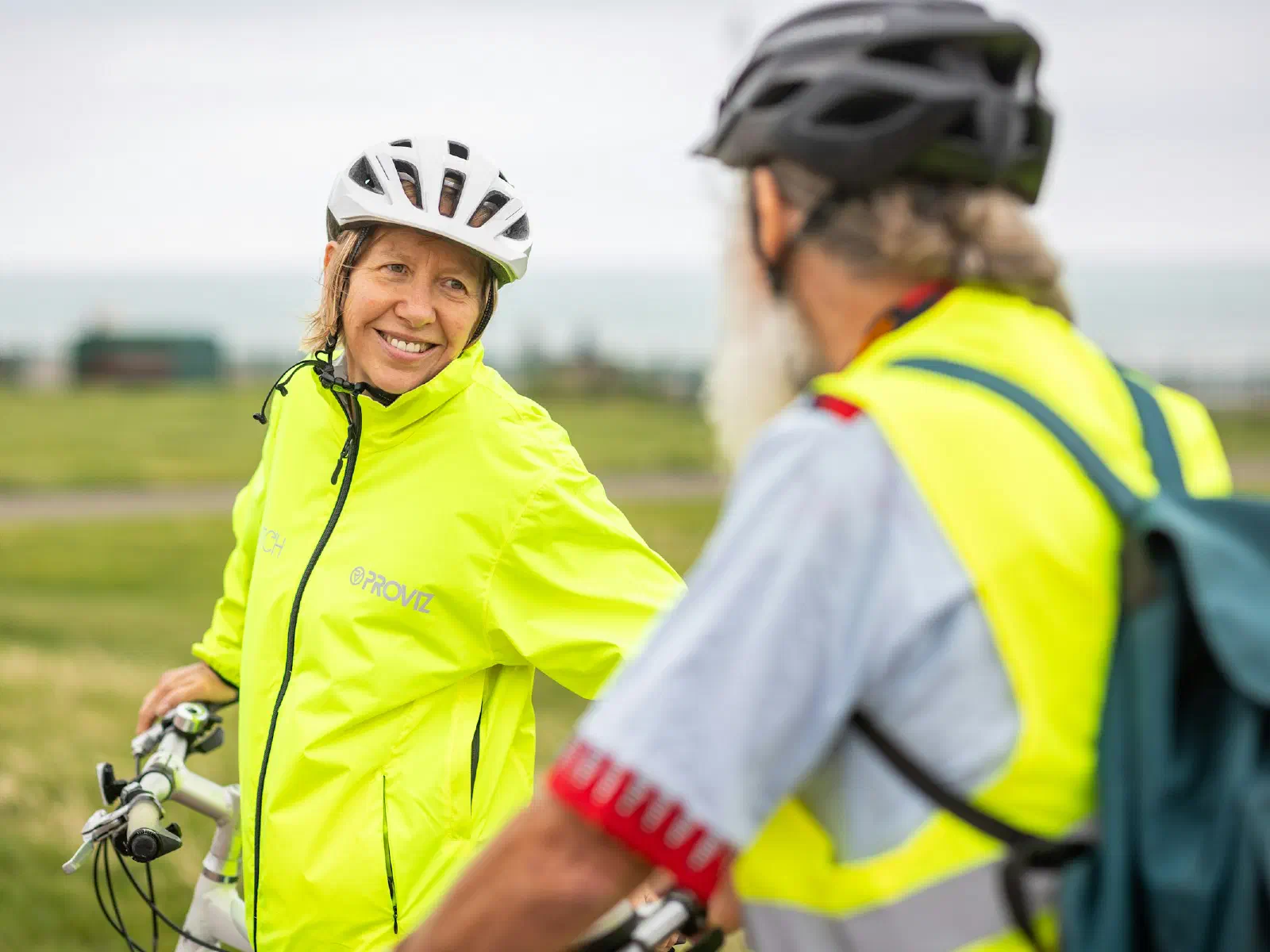 Une femme et un homme casqués, en vestes jaunes, se sourient pendant une pause à vélo près de la mer.