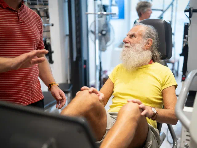 Un homme à barbe blanche fait un exercice de renforcement des jambes sur une machine, en écoutant un encadrant.