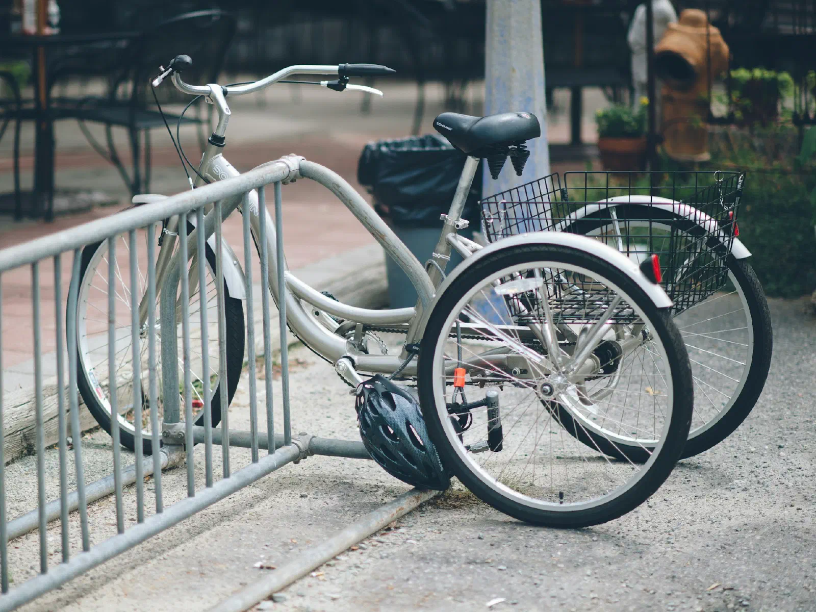 Un tricycle adulte argenté avec panier arrière, garé contre une barrière métallique.