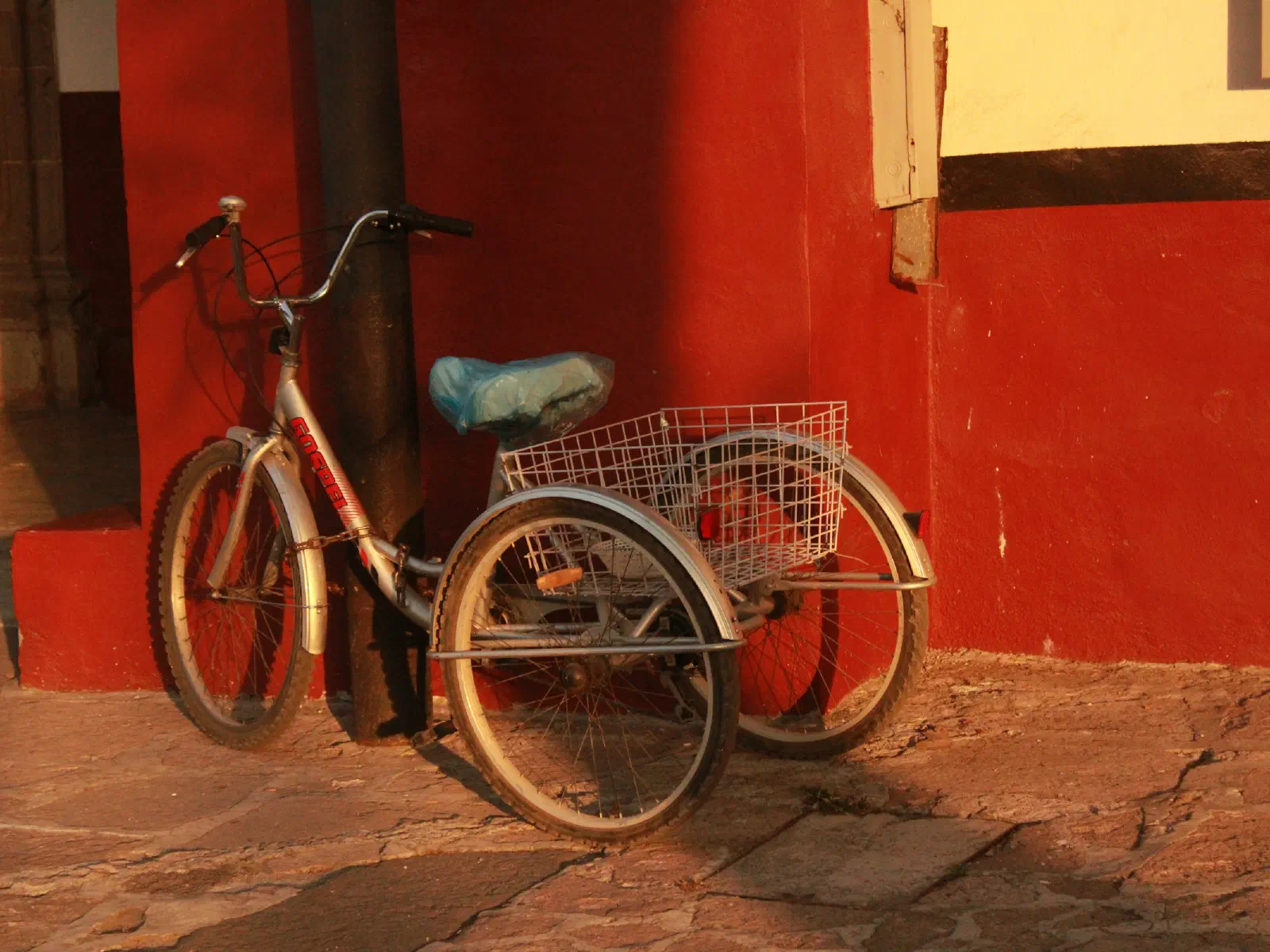 Un tricycle à grand panier arrière garé contre un mur rouge.