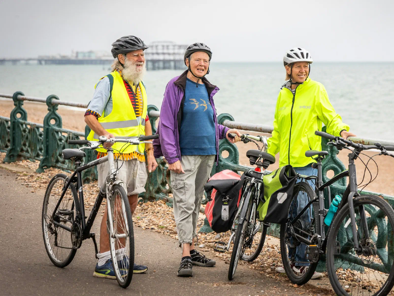 Trois cyclistes aux cheveux blancs, casqués, discutent à côté de leurs vélos sur une promenade en bord de mer.