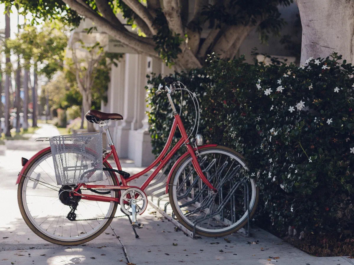 Un vélo rouge à cadre ouvert, avec panier métallique, rangé à un arceau devant une haie fleurie.